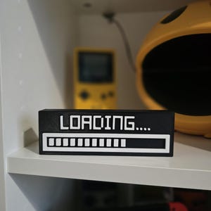 May include: A black rectangular block with the word "LOADING...." in white. Below the text is a white progress bar. The block sits on a white shelf, with a yellow Pac-Man helmet and a yellow handheld game console in the background.