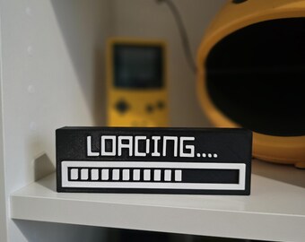 May include: A black and white rectangular block with the word "LOADING...." in white letters. Below the text is a white progress bar. The block is sitting on a white shelf, with a yellow Pac-Man helmet and a yellow handheld game console in the background.