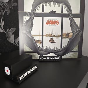 May include: A black and white vinyl record display featuring a shark jaw and the movie poster for "Jaws." A small black sign reads "NOW SPINNING." A black record stand with a red and white circle is also present.