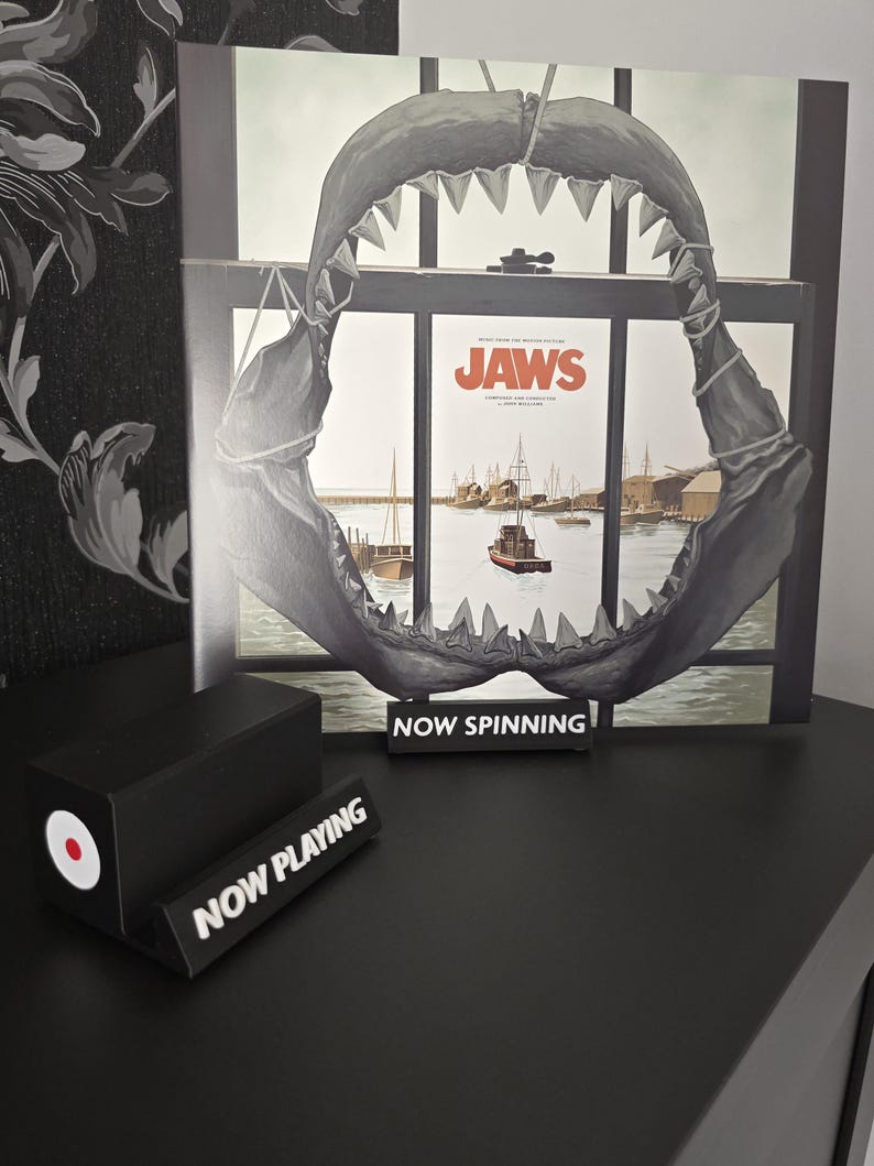 May include: A black and white vinyl record display featuring a shark jaw and the movie poster for "Jaws." A small black sign reads "NOW SPINNING." A black record stand with a red and white circle is also present.
