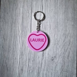 May include: Pink heart-shaped keychain with a silver ring and chain. The heart has a pink outline and the name "Laurie" is written in white inside the heart.