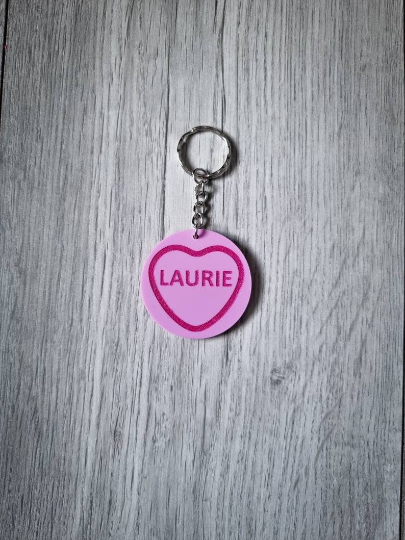May include: Pink heart-shaped keychain with a silver ring and chain. The heart has a pink outline and the name "Laurie" is written in white inside the heart.
