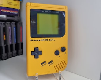 May include: A yellow Nintendo Game Boy handheld video game console with a black screen and buttons. The console is on a clear acrylic stand.