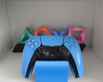 May include: A light blue video game controller rests on a matching blue stand. The controller has black accents and buttons. In the background are colorful geometric shapes, including a green triangle, an orange circle, a blue X, and a pink square.