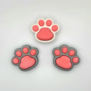 May include: Three paw print shaped keychains. One keychain is white with a pink paw print inside. The other two keychains are gray with a pink paw print inside.
