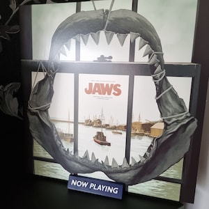 May include: A framed album cover for the movie "Jaws" featuring a shark jaw overlay. The album art depicts a harbor scene. A small blue sign reads "NOW PLAYING".