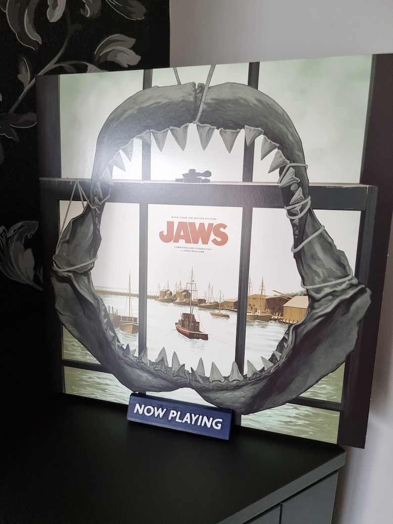 May include: A framed album cover for the movie "Jaws" featuring a shark jaw overlay. The album art depicts a harbor scene. A small blue sign reads "NOW PLAYING".