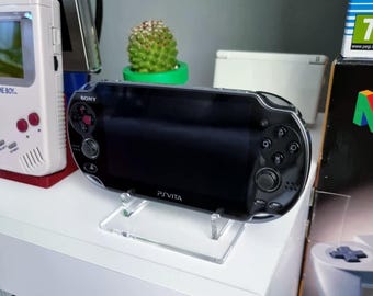 May include: A black Sony PlayStation Vita handheld gaming console with a clear acrylic stand. The console has a black screen and white buttons. The text "PS VITA" is printed on the front of the console.