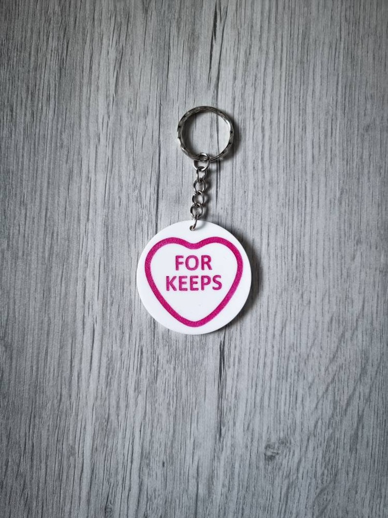 May include: White heart-shaped keychain with a pink outline and the text "FOR KEEPS" in pink letters.