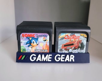 May include: A blue Game Gear game cartridge holder with the words "GAME GEAR" in white. Two black game cartridges are displayed, one featuring Sonic the Hedgehog and the other OutRun. The holder is on a white shelf.