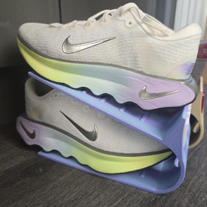 May include: White athletic trainers with a silver swoosh logo, on a two-tier shoe rack. The rack has a gradient of colours, from yellow to purple. The trainers are designed for running.