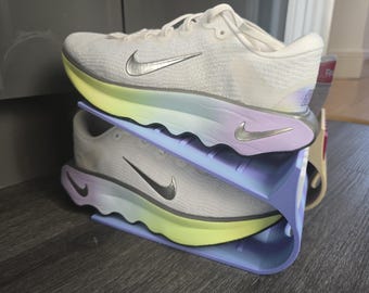 May include: White athletic shoes with a silver swoosh logo, resting on a two-tiered shoe rack. The rack has a gradient of colors, transitioning from yellow to purple. The shoes are designed for running.