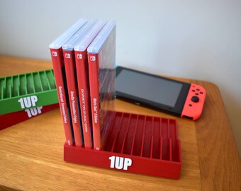 May include: Red and green plastic game cartridge storage stands with the text "1UP" printed on the front. The stands are holding several video game cartridges in their slots. A Nintendo Switch console and controller are also visible on the table.