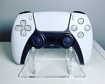 May include: A white and black video game controller with a clear acrylic stand. The controller has a white face with black buttons and a black back.