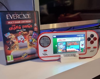 May include: A red and white Evercade handheld gaming console with a white stand. The screen shows a game called "The Oliver Twins Collection" with a cartoon character on the screen. The console has a red button, a gray directional pad, and a black and white button labeled "Select".