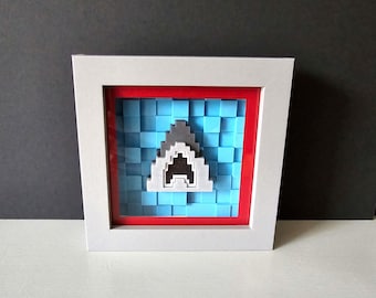 May include: A framed pixel art piece depicting a shark fin in a blue and white pixelated background. The frame is white with a red inner border.