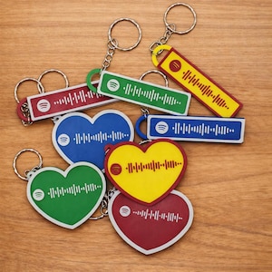 May include: A selection of keyrings in various shapes and colours, including hearts and rectangles. Each keyring features a Spotify logo and a unique audio waveform design. The keyrings are attached to silver key rings and are arranged on a wooden surface.