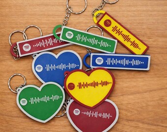 May include: A collection of keychains in various shapes and colors, including hearts and rectangles. Each keychain features a Spotify logo and a unique audio waveform design. The keychains are attached to silver key rings and are arranged on a wooden surface.
