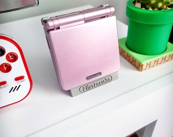 May include: A pink Game Boy Advance SP handheld gaming console with a Nintendo logo on the front. The console is sitting on a white surface with a green and brown potted cactus in the background.
