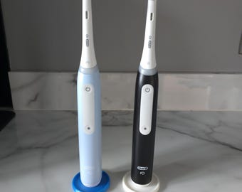 May include: Two electric toothbrushes, one light blue and one black, with white accents and blue brush heads. Each toothbrush has a power button and is displayed on a stand. The Oral-B IO logo is visible on the black toothbrush.