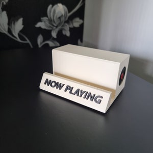 May include: A cream-colored vinyl record holder with the words "NOW PLAYING" in black lettering. The holder has a rectangular shape with a circular red accent on the side. The holder is on a black surface.