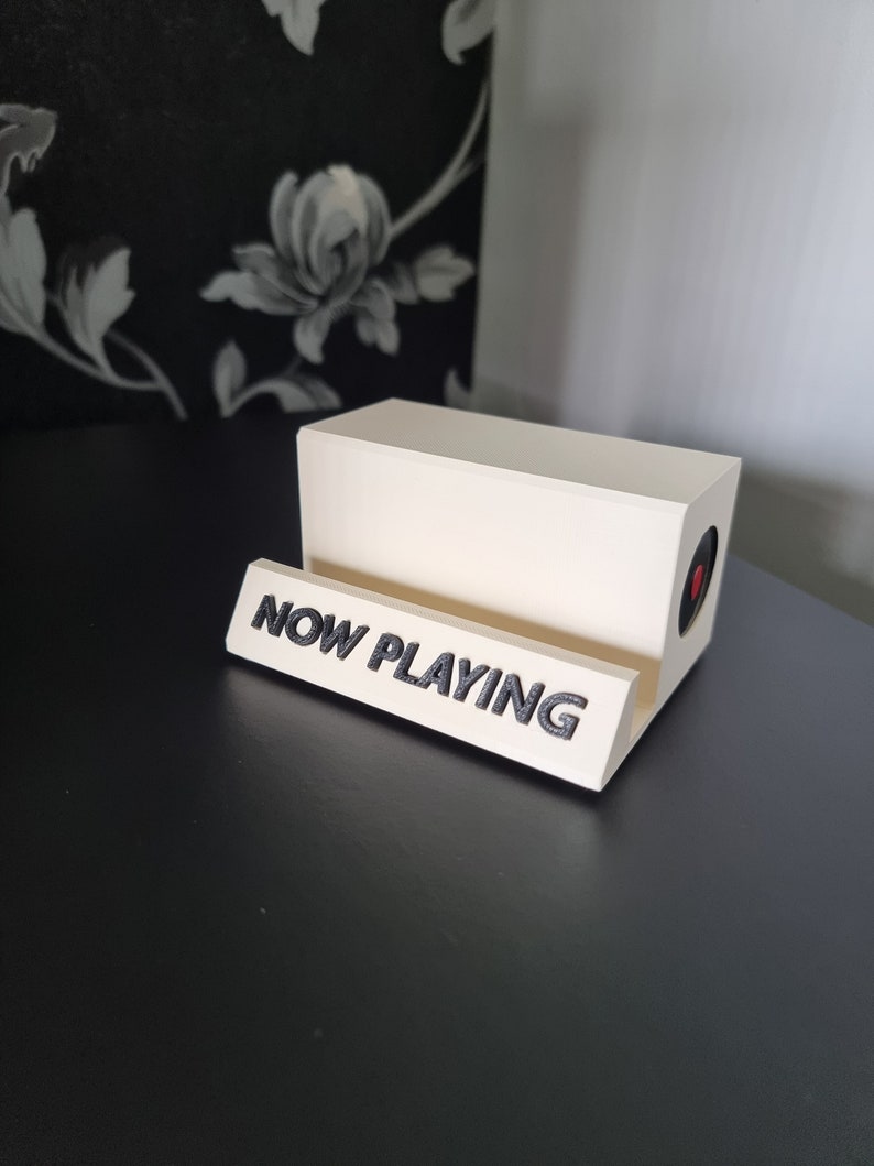 May include: A cream-colored vinyl record holder with the words "NOW PLAYING" in black lettering. The holder has a rectangular shape with a circular red accent on the side. The holder is on a black surface.