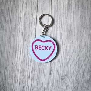 May include: A white heart-shaped keychain with a pink outline and the name "Becky" written in pink inside. The keychain is attached to a silver metal ring.