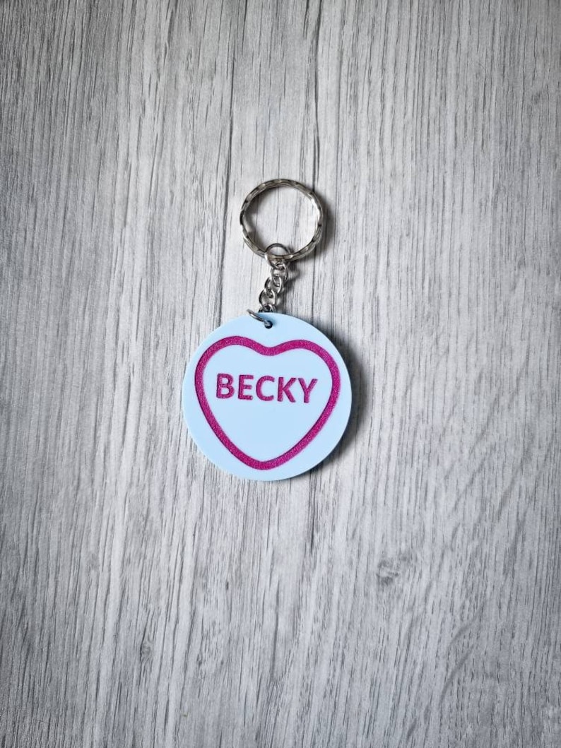 May include: A white heart-shaped keychain with a pink outline and the name "Becky" written in pink inside. The keychain is attached to a silver metal ring.