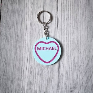 May include: A light blue heart-shaped keychain with a pink outline and the name "MICHAEL" written in pink inside. The keychain is attached to a silver key ring.