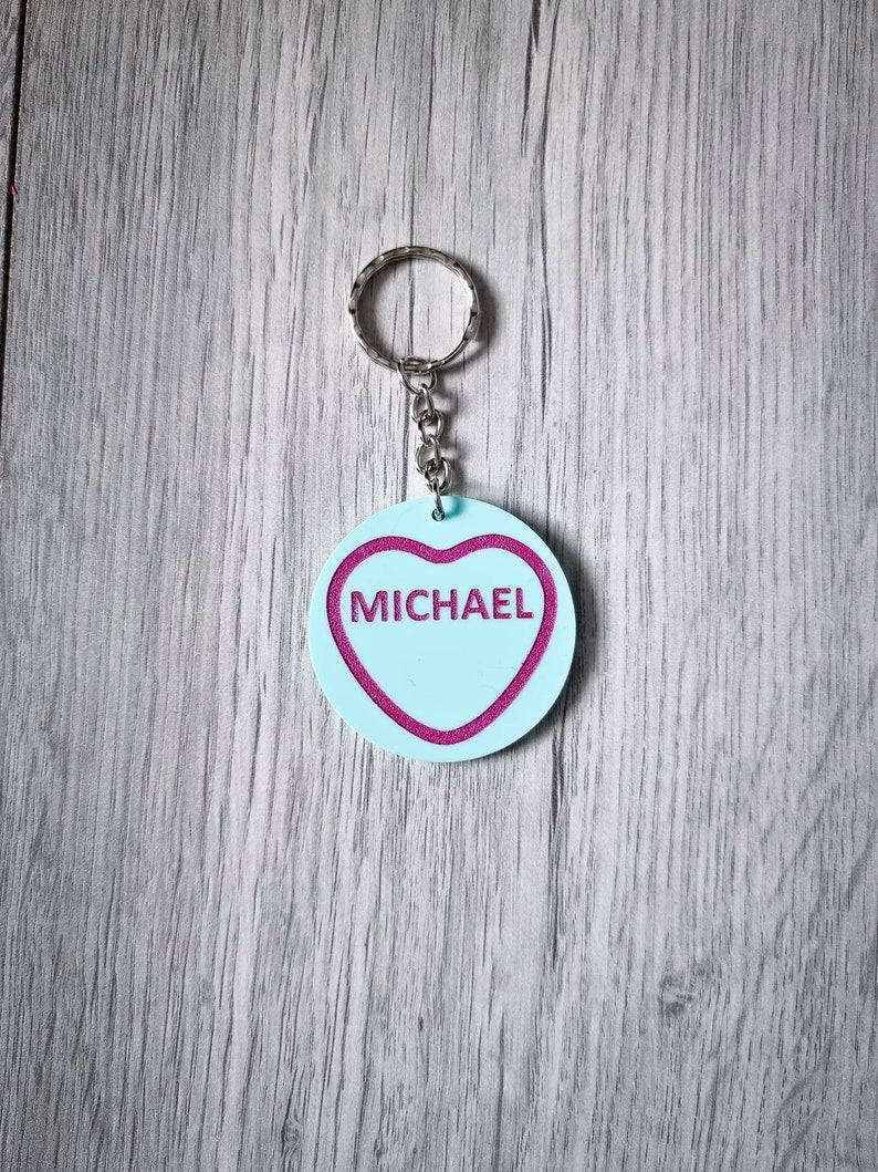May include: A light blue heart-shaped keychain with a pink outline and the name "MICHAEL" written in pink inside. The keychain is attached to a silver key ring.