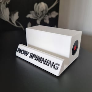 May include: White 3D printed record player stand with the words "NOW SPINNING" on the front. The stand has a black circle with a red dot in the centre.