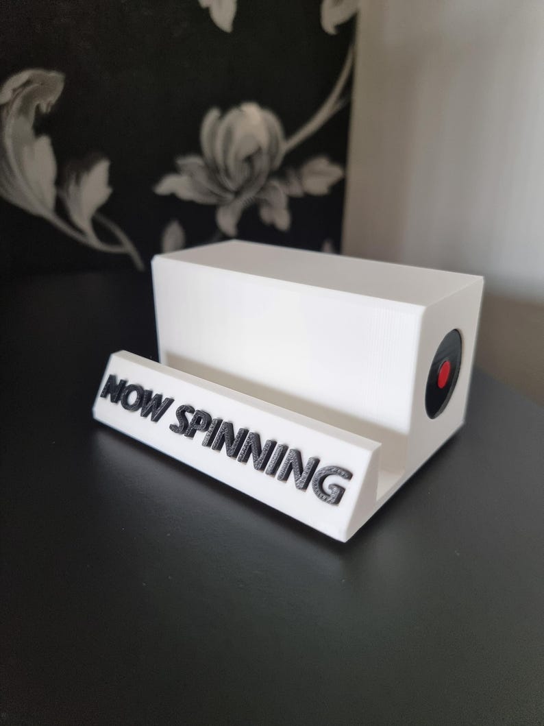 May include: A white vinyl record holder with a black and red accent. The holder has a rectangular shape with a slot for a record and the words "NOW SPINNING" in black lettering.