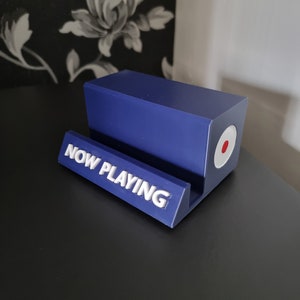 May include: A blue rectangular phone stand with the words "NOW PLAYING" in white lettering. The stand has a circular design with red and white accents on the side. The stand is on a black surface.