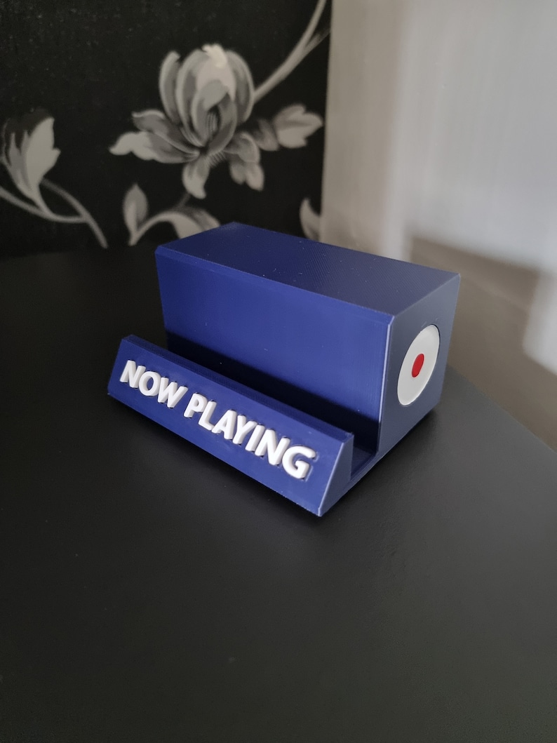 May include: A blue rectangular phone stand with the words "NOW PLAYING" in white lettering. The stand has a circular design with red and white accents on the side. The stand is on a black surface.