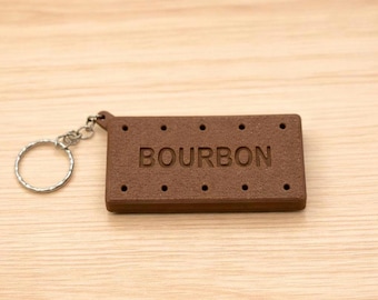 May include: A brown keychain shaped like a biscuit, with the word "BOURBON" engraved on it. The keychain has a silver ring attached. The biscuit has small, evenly spaced holes, and the overall design is simple and food-themed.