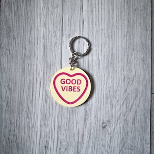 May include: A yellow heart-shaped keychain with a silver ring. The heart has a pink outline and the words "GOOD VIBES" printed in pink.