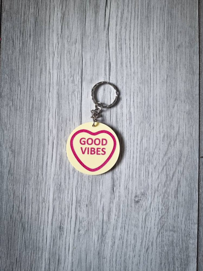 May include: A yellow heart-shaped keychain with a silver ring. The heart has a pink outline and the words "GOOD VIBES" printed in pink.
