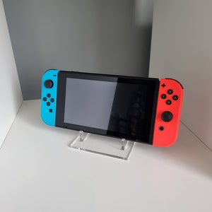 May include: A black handheld gaming console with blue and red controllers is displayed on a clear acrylic stand. The console is resting on a white shelf. The controllers have buttons labeled with letters and symbols.