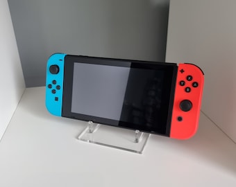 May include: A black handheld gaming console with blue and red controllers is displayed on a clear acrylic stand. The console is resting on a white shelf. The controllers have buttons labeled with letters and symbols.