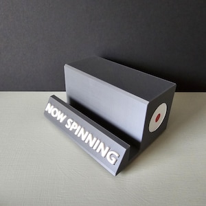 May include: A gray vinyl record stand with the text "NOW SPINNING" in white. The stand has a rectangular base and a slot to hold a record. A white circle with a red dot is on the side.