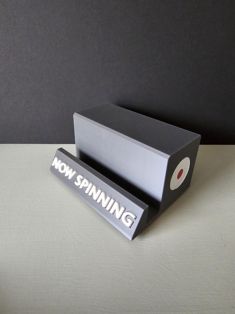 May include: A gray vinyl record stand with the text "NOW SPINNING" in white. The stand has a rectangular base and a slot to hold a record. A white circle with a red dot is on the side.