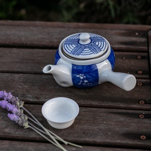 Può includere: Una teiera in ceramica bianca e blu con coperchio abbinato e una piccola tazza bianca. La teiera presenta un motivo geometrico blu e bianco e un manico. Un rametto di lavanda è accanto alla tazza su una superficie di legno.
