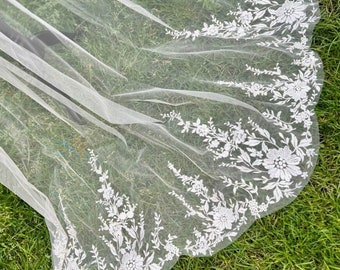 May include: A white tulle veil with a scalloped edge and floral embroidery. The veil is laid out on a green grassy surface.