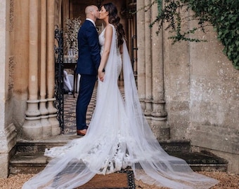 May include: A bride and groom kiss in front of a stone archway. The bride is wearing a white wedding dress with a long veil and the groom is wearing a dark suit.