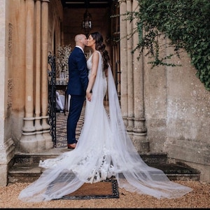 May include: A bride and groom kiss in front of a stone archway. The bride is wearing a white wedding dress with a long veil and the groom is wearing a dark suit.