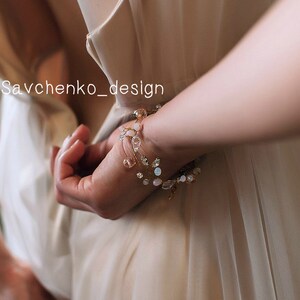 May include: A delicate gold bracelet with pink and white beads, featuring a vine-like design. The bracelet is worn on a person's wrist, with the hand holding a white fabric.