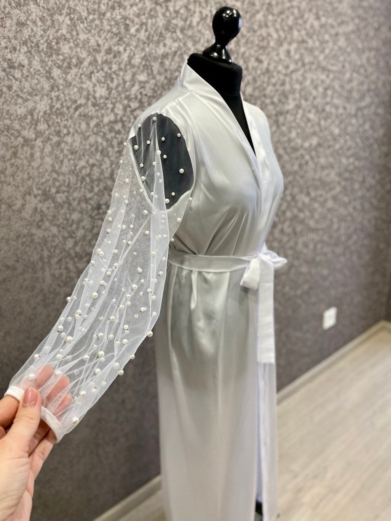 May include: White satin robe with long sleeves featuring a sheer mesh overlay adorned with pearl embellishments.