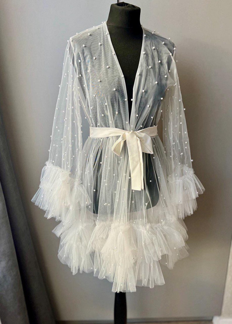 May include: A white sheer robe with a satin sash and pearl embellishments. The robe has a V-neckline and long sleeves with ruffles at the cuffs.