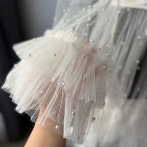 May include: White tulle fabric with a layered ruffle detail and pearl embellishments. The fabric is sheer and delicate, with a soft, flowing texture.