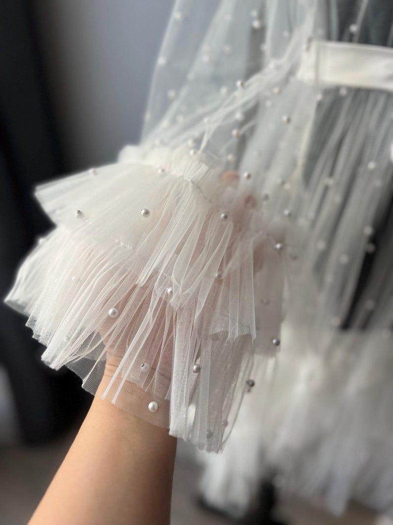 May include: White tulle fabric with a layered ruffle detail and pearl embellishments. The fabric is sheer and delicate, with a soft, flowing texture.